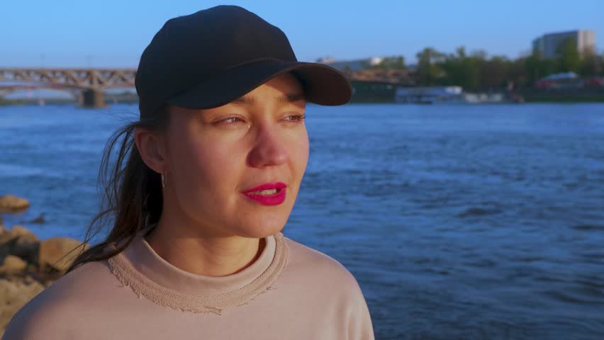 Portrait of a young woman in a baseball cap standing near the river at sunset.