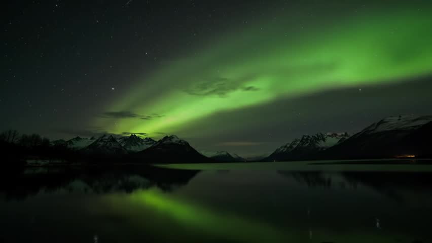 A stunning view of the Aurora Borealis (Northern Lights) over snowy mountains and a tranquil lake. The bright green light arcs dramatically across the starry night sky.