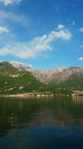 Wide landscape of Lake Como Italy with deep blue water and majestic green mountain peaks under bright summer sky with soft white clouds
