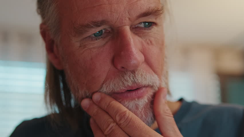 A close-up shot captures a thoughtful senior man with a beard as his expression transforms into a warm, genuine smile. His blue eyes convey happiness and contentment in a comfortable, home setting.