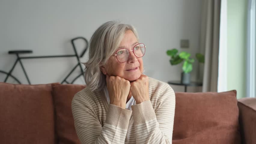 Peaceful senior woman with grey hair and glasses relaxing on the couch at home, looking out the window with a happy and pensive expression, enjoying her retirement and reminiscing