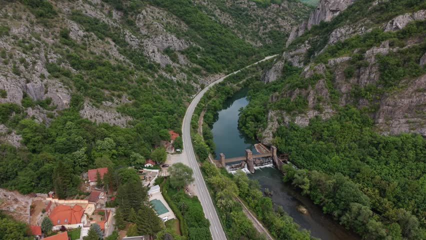 A view shows a winding road by a river surrounded by tall green mountains and a dam in the valley.