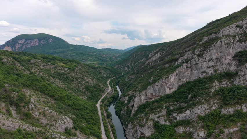 A river flows through a valley surrounded by tall mountains and green trees. The sky is partly cloudy and the landscape is wide and open.