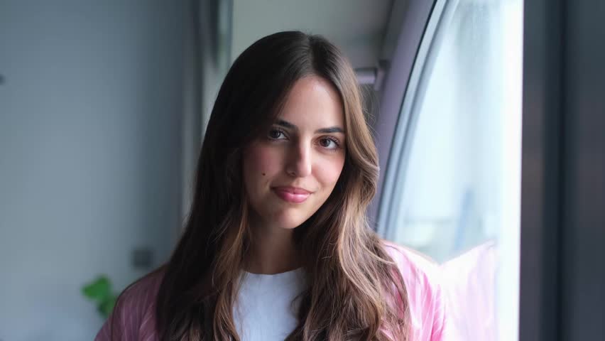 Portrait of a beautiful young caucasian woman with long brown hair smiling genuinely at the camera while standing by a window, with soft natural light illuminating her happy face