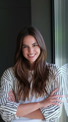 Cheerful young brunette woman standing by a window with her arms crossed. This beautiful female entrepreneur is looking away, smiling confidently and feeling optimistic about her future
