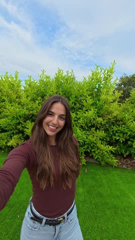 Cheerful smiling woman holding a camera, filming herself for a social media vlog, greeting her followers with a wave, and showing a peace sign while standing outdoors in a lush green garden