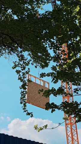 Timber wall element flying through air on metal hook behind tree branches. Eco friendly building materials transport at sunny outdoor site