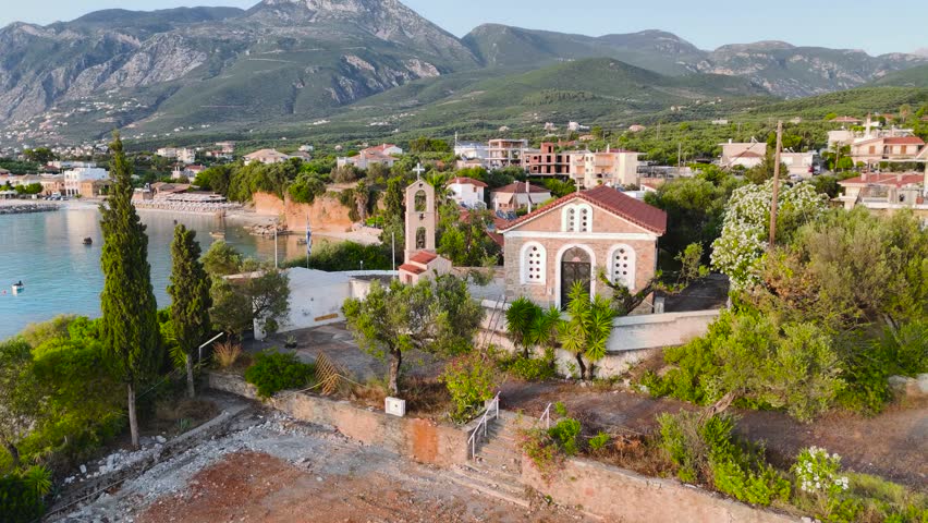 Cinematic aerial drone footage featuring a small Greek seaside church standing on a rocky coastal slope near Kalamata in the Peloponnese, Greece. The scene includes turquoise Mediterranean water, a quiet coastal village, rocky shoreline, small bay and dramatic mountain backdrop in soft golden evening light. Ideal for travel videos, tourism content, religion and spirituality themes, Mediterranean lifestyle projects, scenic destination backgrounds and coastal aerial sequences.