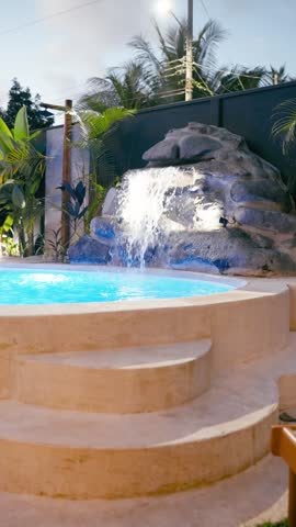 Sunny resort hotel swimming pool with an artificial waterfall flowing into turquoise water. Beautiful tropical plants surround the private spa area