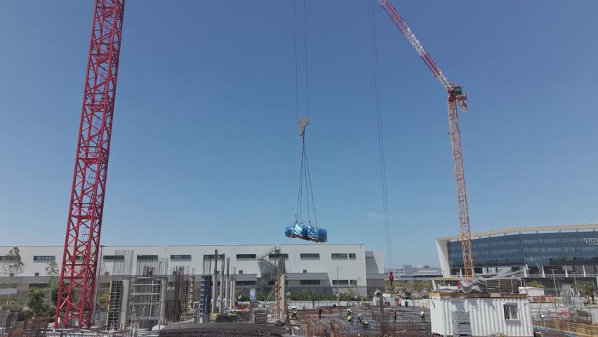 Two large cranes lifting a heavy load at an active israeli construction site with industrial buildings and blue sky background
