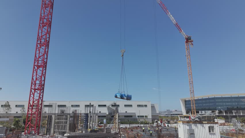 Two large cranes lifting a blue wrapped heavy load over an active israeli construction site with industrial buildings under clear blue sky
