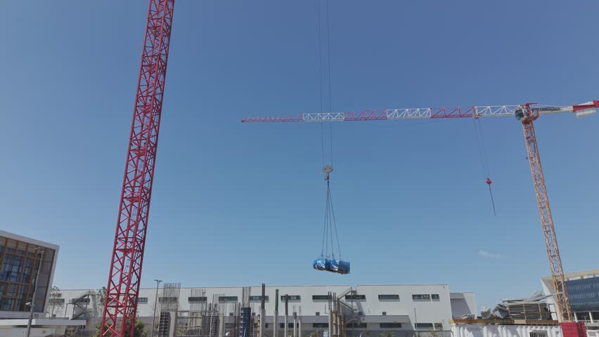 Two large cranes lifting a heavy load at an active israeli construction site with industrial buildings and blue sky background