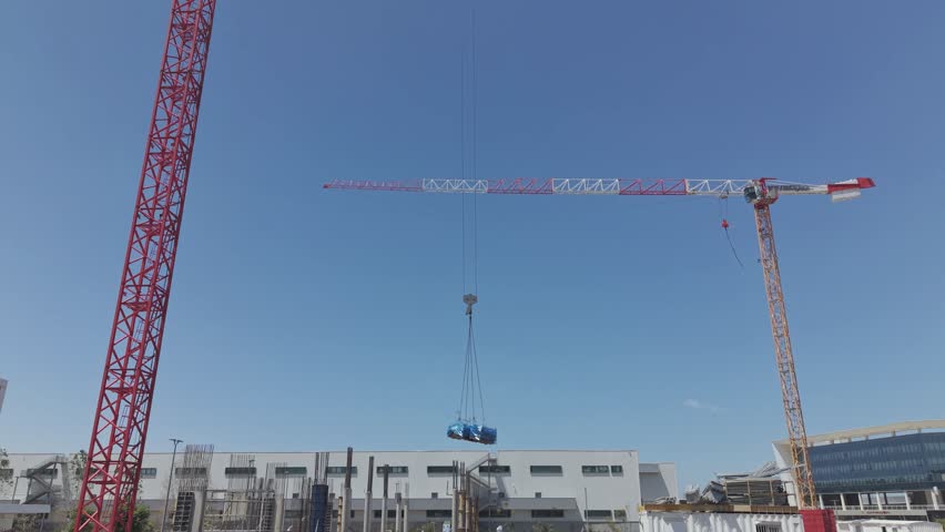 Two large cranes lifting a blue wrapped load at an israeli construction site with industrial buildings against clear blue sky
