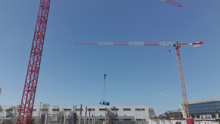 Two large red cranes lifting a blue wrapped load against clear blue sky at an israeli industrial construction site

