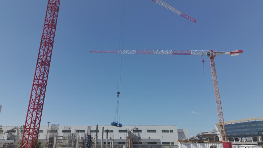 Tower crane lifting heavy blue load against clear blue sky at urban construction site with two cranes
