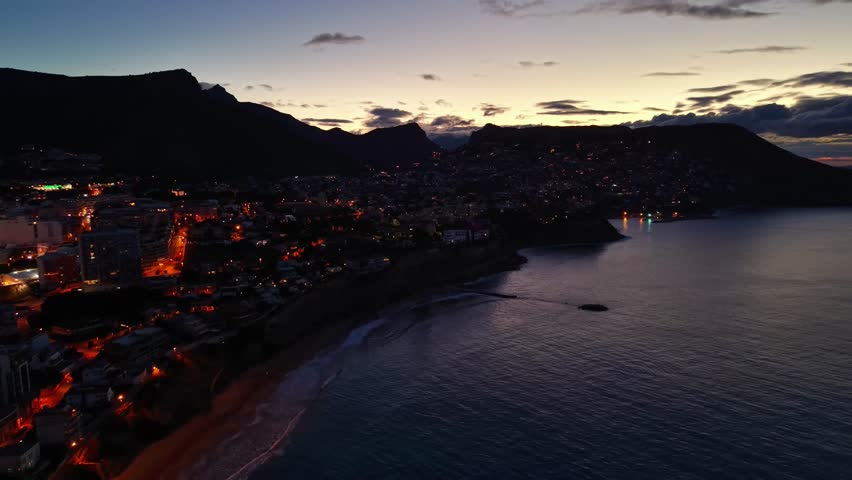 Aerial coastal town at dusk illuminated by streetlights, bay curve with dark silhouette mountains, glowing