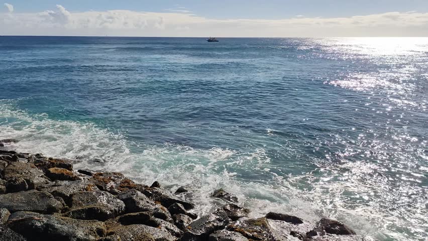Honolulu, USA - The image showcases ocean waves crashing over dark rocks along the coastline, with a distant boat on the horizon and glimmering water.