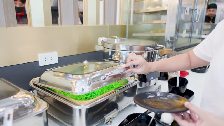 A person opens a stainless steel chafing dish and serves green noodles at a buffet
