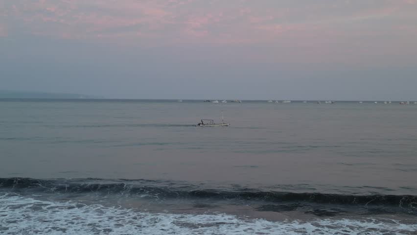 A traditional jukung sails past in the distance at dusk as fishermen return home from the sea.