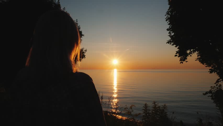 Silhouette of woman gazing at beautiful sunset over calm sea. Golden sunlight reflects on water. Peaceful nature scene, cinematic footage.