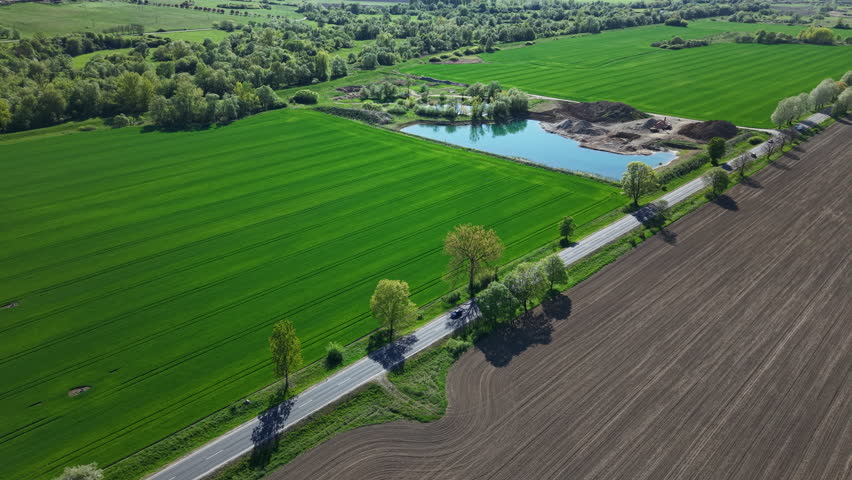 An aerial perspective shows a road dissecting vibrant green fields and ploughed soil with a small blue pond nearby.