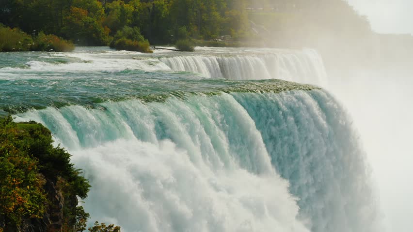 Powerful Niagara falls cascading over rocky precipice with dense water mist rising from turbulent river basin at base. Scenic natural landscape featuring rushing currents and splashing white foam water.