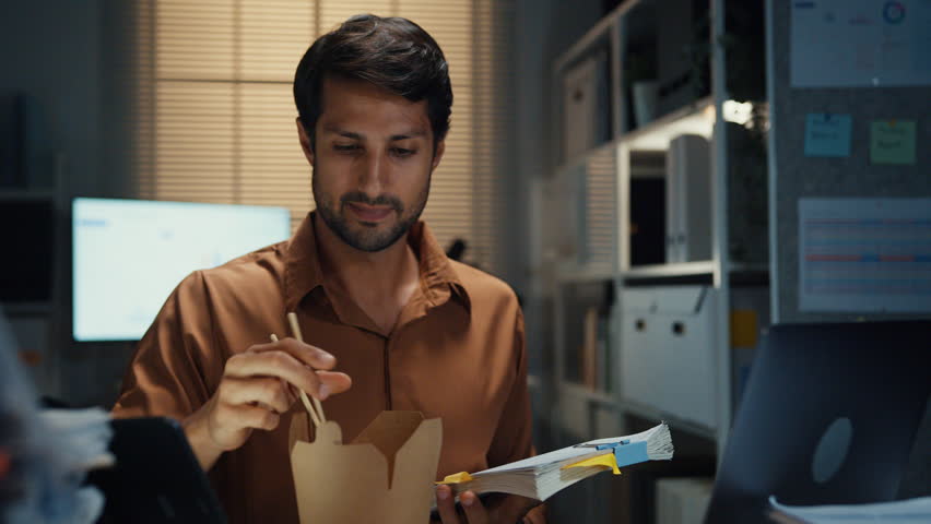 Young Latin businessman eating takeout instant noodles and reviewing document on laptop at desk in dark night office. Overtime working late, overworked deadline, fast food, corporate problem concept.