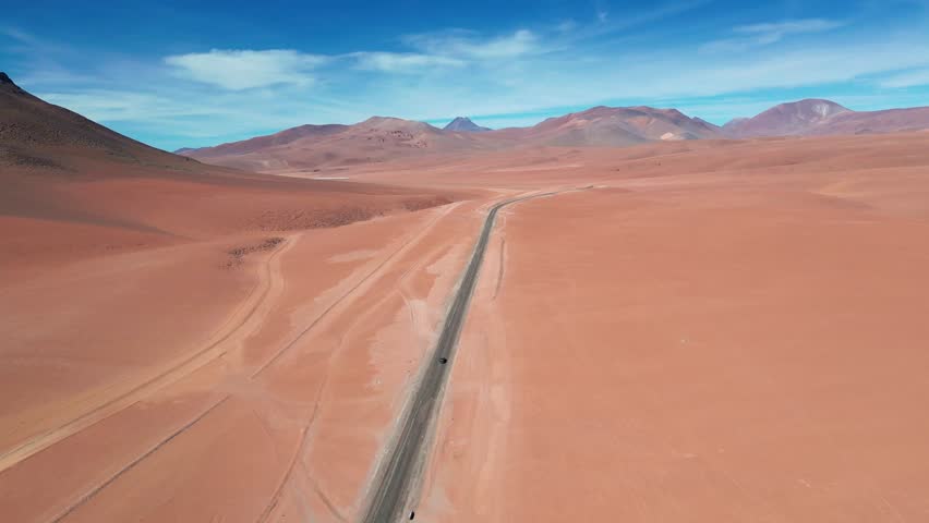 Tracking drone shot following a vehicle along a straight highway cutting through vast red sand desert framed by volcanic hills and mountain range. Atacama Desert, Chile