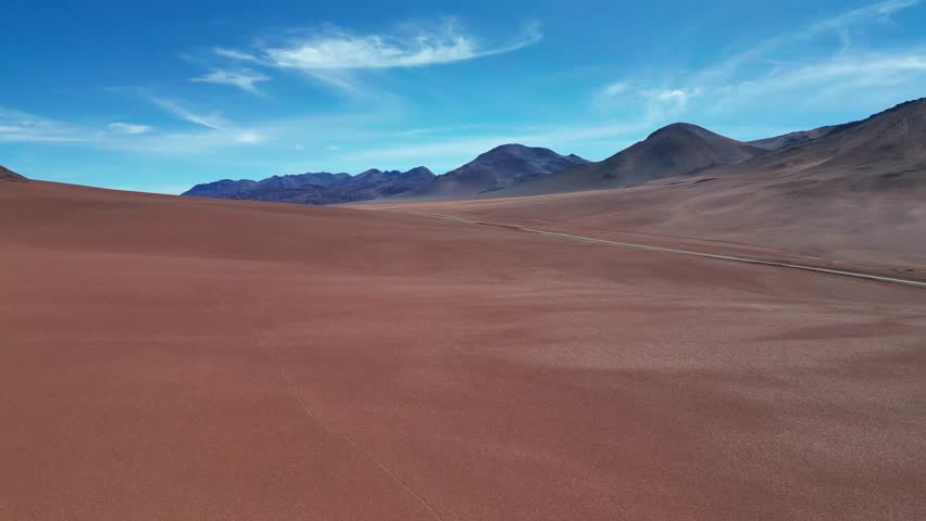 Ascending drone shot rising over a pristine vast red sand desert plain revealing a chain of volcanic cones stretching across the horizon under vivid blue sky. Atacama Desert, Chile