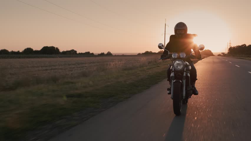 Frontal view of man wearing helmet riding classic motorcycle on asphalt road at golden hour. Warm sunlight glares behind rider driving through rural landscape. Cinematic motion footage of road trip.