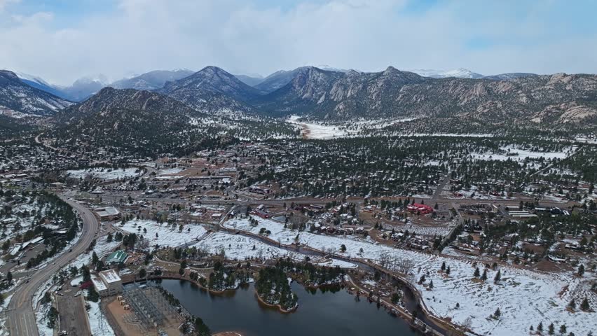 Estes Park town with lake and surrounding forest below snow capped Rocky Mountains, aerial drone overview