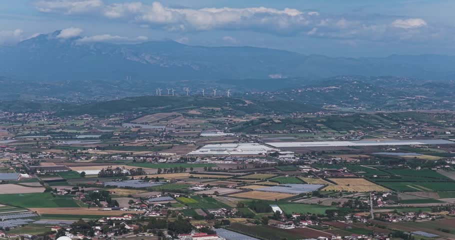 Aerial view of the vast agricultural landscape with greenhouses, rural settlements, and wind turbines on the distant ridge under a cloudy sky in Capaccio, Campania, Italy.