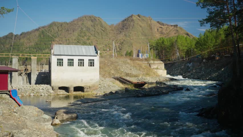 Power Plant Nearby River. Hydropower Installation Located Beside Lively Mountain River Terrain. Facility Harnessing Energy From Flowing Mountain Stream Amidst Rugged Landscape Under Clear Sky
