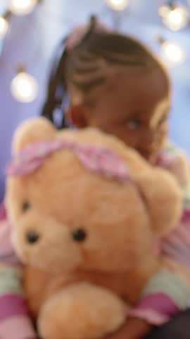 Vertical video: Hugging tan teddy, braided girl in striped top in bedroom reacting to offscreen cue. Bear, plush, child, pastel, cozy, lights, vertical video