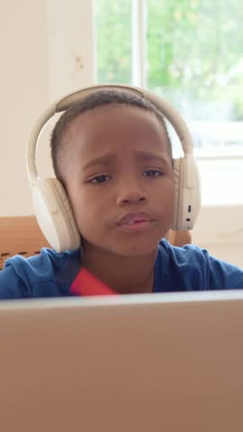 Vertical video: Listening boy wearing white headset blue T to laptop audio at home writing notes. Headphones, concentration, notetaking, notebook, pencil, desk, vertical video