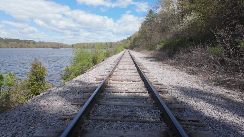 Walking along train tracks in a POV view. The tracks are surrounded by water on one side and trees and foliage on the other side on a beautiful day in nature.