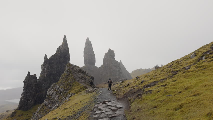 The Storr, Scotland, a person is hiking on narrow path leading toward rocky peaks with misty atmosphere in Scottish highlands