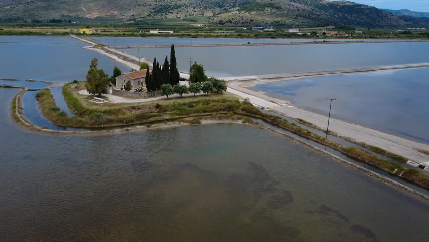 Aerial close view above a chapel island inside shallow salt flats with calm water, trees and traditional Mediterranean architecture in Greece, Europe.