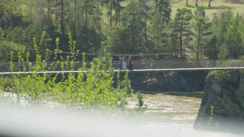 Stream Crossing With Forest Backdrop. Travelers Traverse River Via Suspension Pathway Amidst Lush Greenery. Hikers Crossing River On Suspension Bridge Near Woodland Scene With Flowing Water