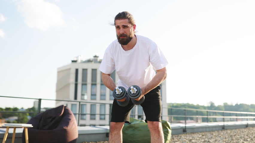 A bearded man with long hair tied back is performing a dumbbell exercise on a rooftop with a modern building in the background