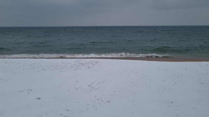 Winter sea with falling snow and waves on sandy beach. Cold coastal landscape with overcast sky and snow-covered shore