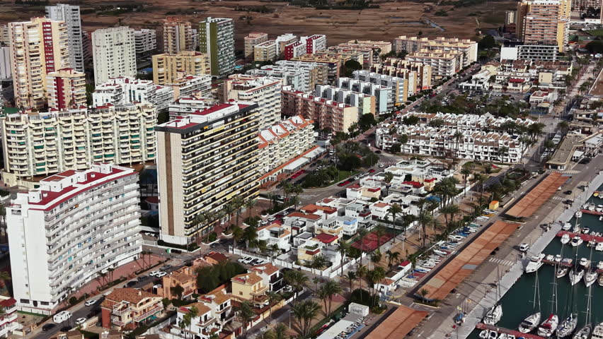 A coastal town with several tall buildings and small houses. Yachts are docked in the marina. Streets run alongside buildings and palm trees.
