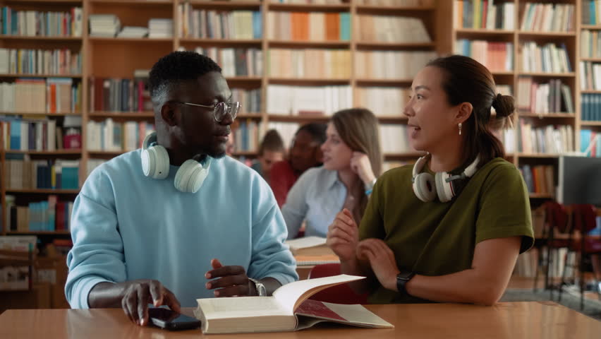 African male student and Asian female student giving high five during collaborative studying in university library with classmates in background.	
