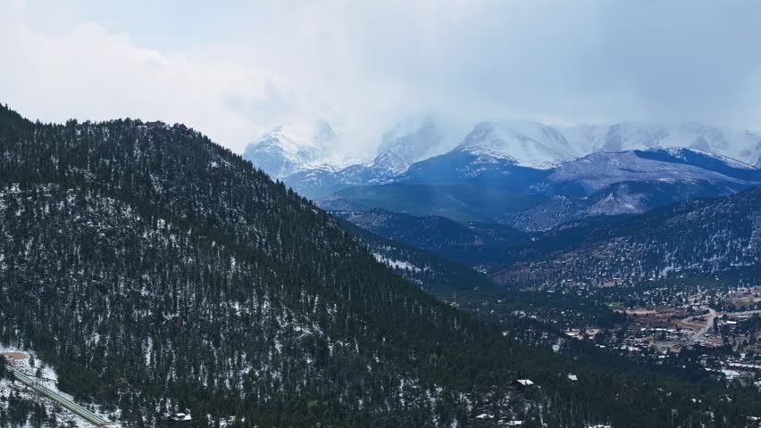 Estes Park valley with forested slopes and snow covered Rocky Mountains under cloudy sky, aerial drone overview