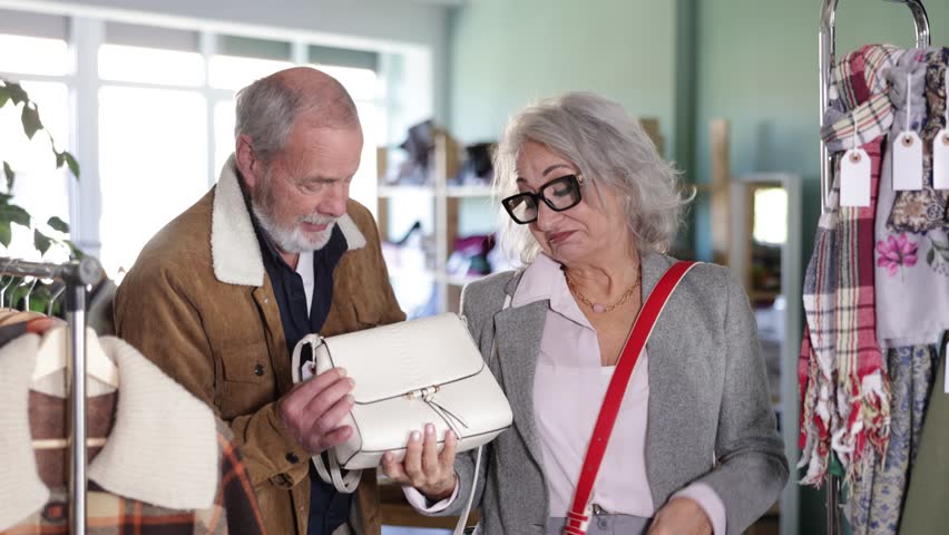 Elderly couple chooses a stylish handbag together, the husband helps his wife with the choice against the background of the shelves with things in the store. High quality 4k footage