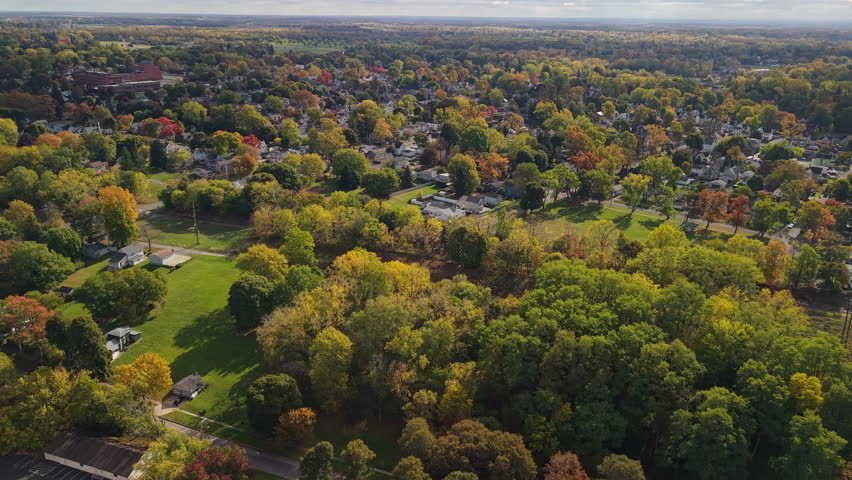 Aerial view of suburban town in USA with residential neighborhoods, green parks and colorful autumn forest under sunny sky.