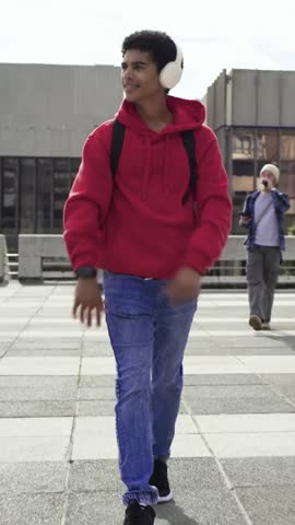 Vertical video: Walking across plaza, man in red hoodie with backpack and headphones checking watch. Urban, pedestrian, streetwear, wearable, movement, daylight, vertical video