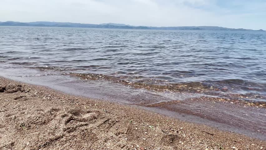 A scenic view of a calm lake, sandy shore, and gently lapping shoreline waves.