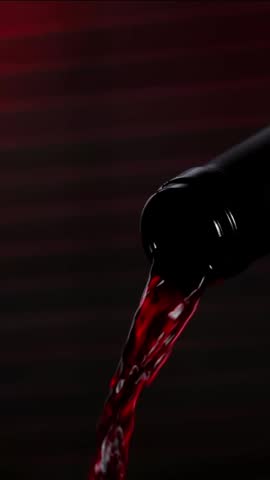 A bottle of red wine is tilted to pour wine into a clear glass. The dark background adds focus to the flowing wine. This takes place in the evening during a dinner gathering.