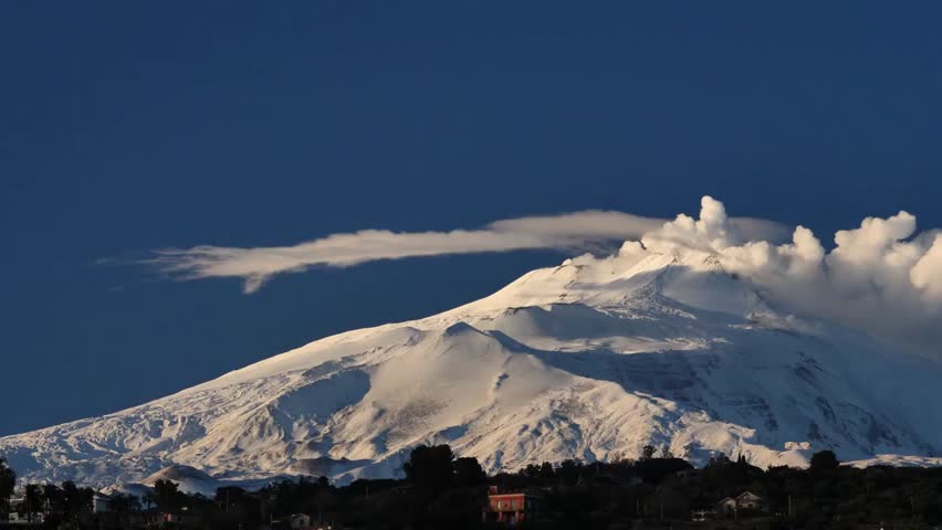 A breathtaking view of Mount Etna, an active stratovolcano, with its summit covered in snow and illuminated by the warm glow of the setting sun.
